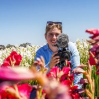 Wide shot of a reporter behind a camera in a field of flowers